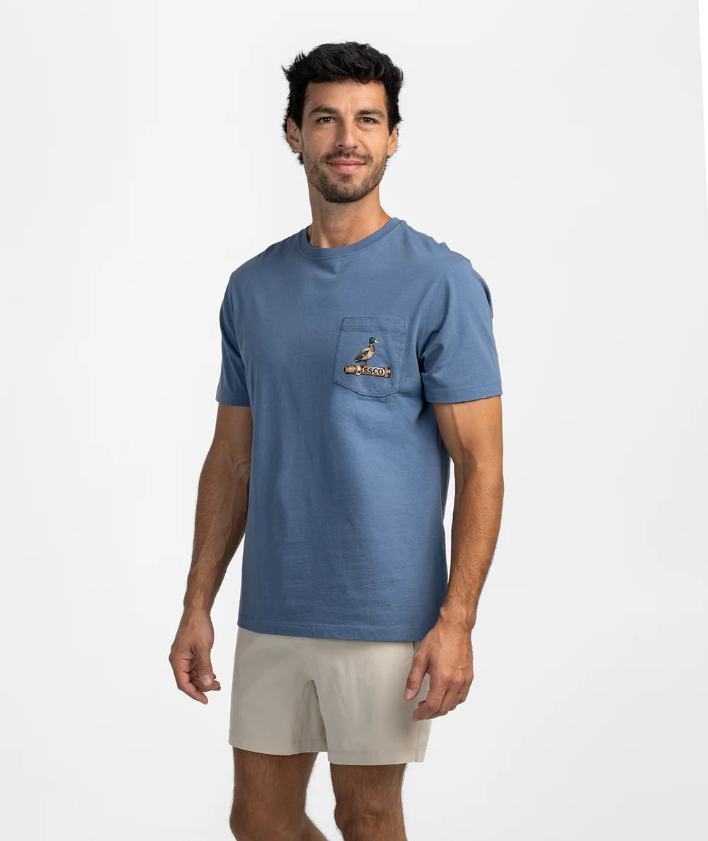 A man modeling a blue crewneck pocket t-shirt with a small embroidered bird logo on the chest pocket, paired with beige shorts, against a light gray studio background.