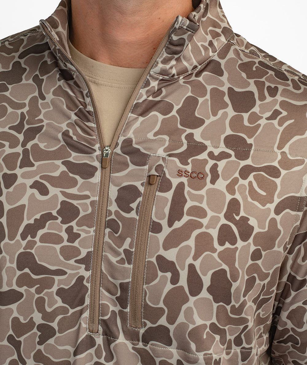 Close-up of a person wearing a brown and beige camouflage patterned quarter-zip pullover with the SSCO logo on the chest pocket.