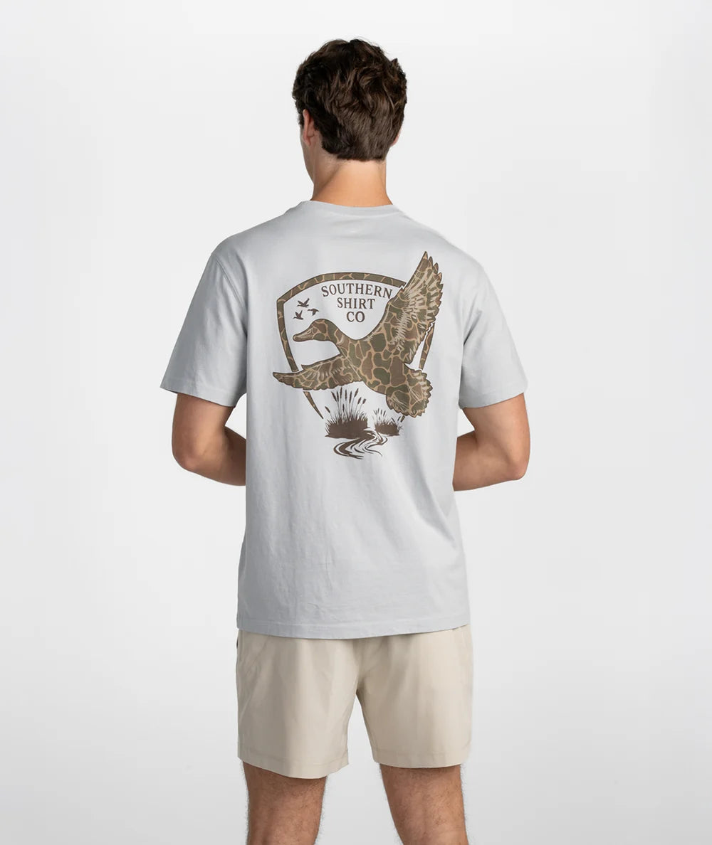 A man modeling a light gray short-sleeve t-shirt with a small brown chest graphic and the brand name 'SSCO' printed on the pocket, paired with beige shorts against a plain white studio background.