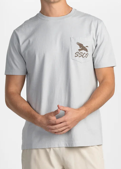A man modeling a light gray short-sleeve t-shirt with a small brown chest graphic and the brand name 'SSCO' printed on the pocket, paired with beige shorts against a plain white studio background.