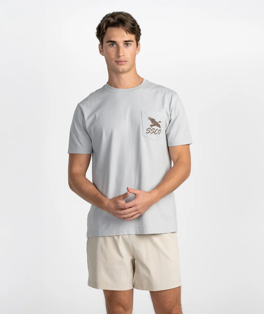 A man modeling a light gray short-sleeve t-shirt with a small brown chest graphic and the brand name 'SSCO' printed on the pocket, paired with beige shorts against a plain white studio background.