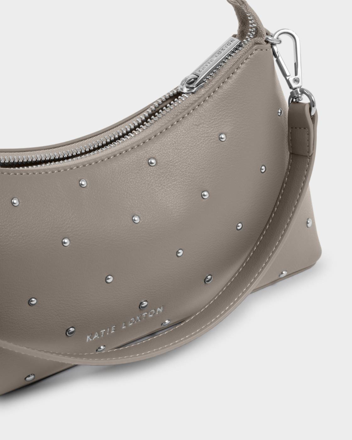 A close-up of a taupe leather shoulder bag by Katie Loxton, decorated with silver studs and featuring a silver zipper and strap.