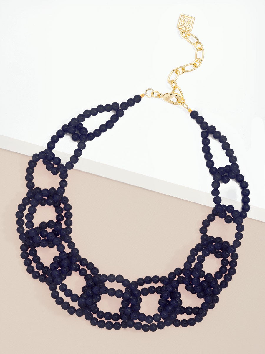 A multi-strand beaded collar necklace in dark navy blue with a gold-tone chain extender and clasp, displayed on a split beige and white background.
