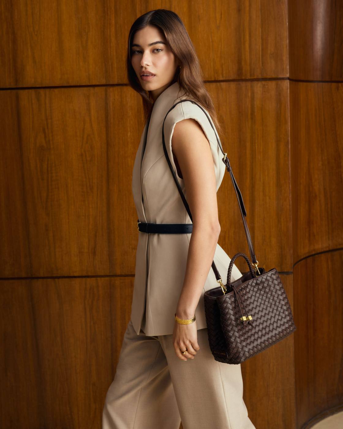 A woman in a beige sleeveless vest and matching trousers carries a dark brown woven leather handbag with gold hardware against a warm wood-paneled background.