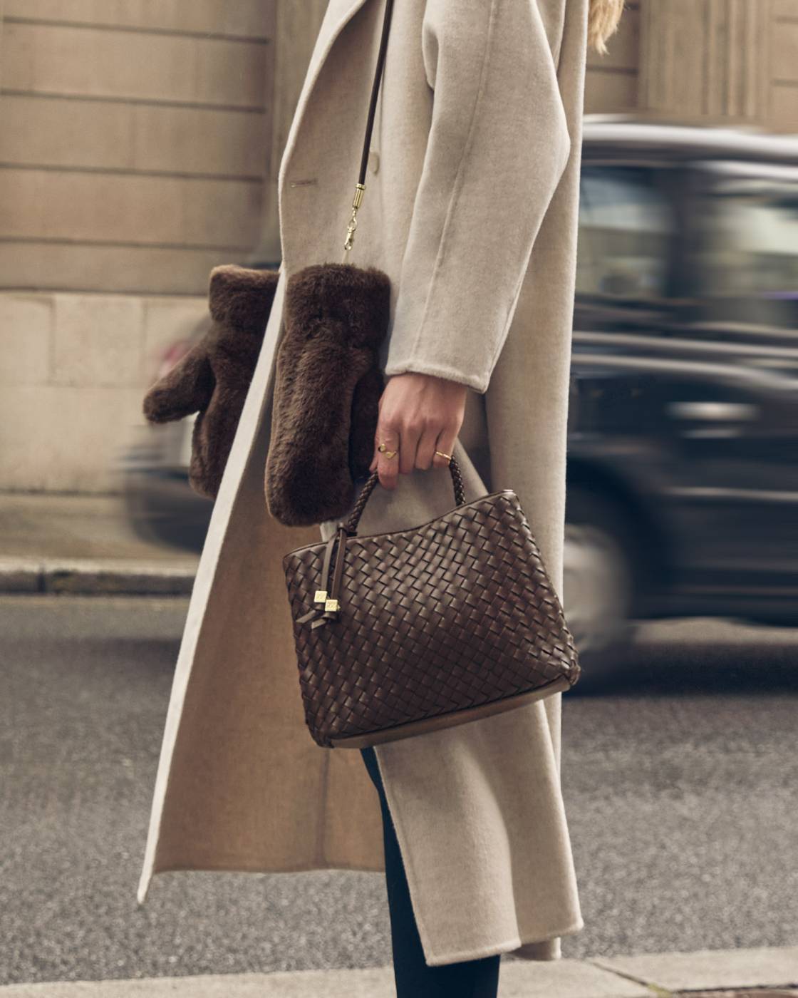 A person in a long beige coat carries a brown woven leather handbag with dark brown faux-fur trim and a matching brown fur accessory, photographed on a city street.
