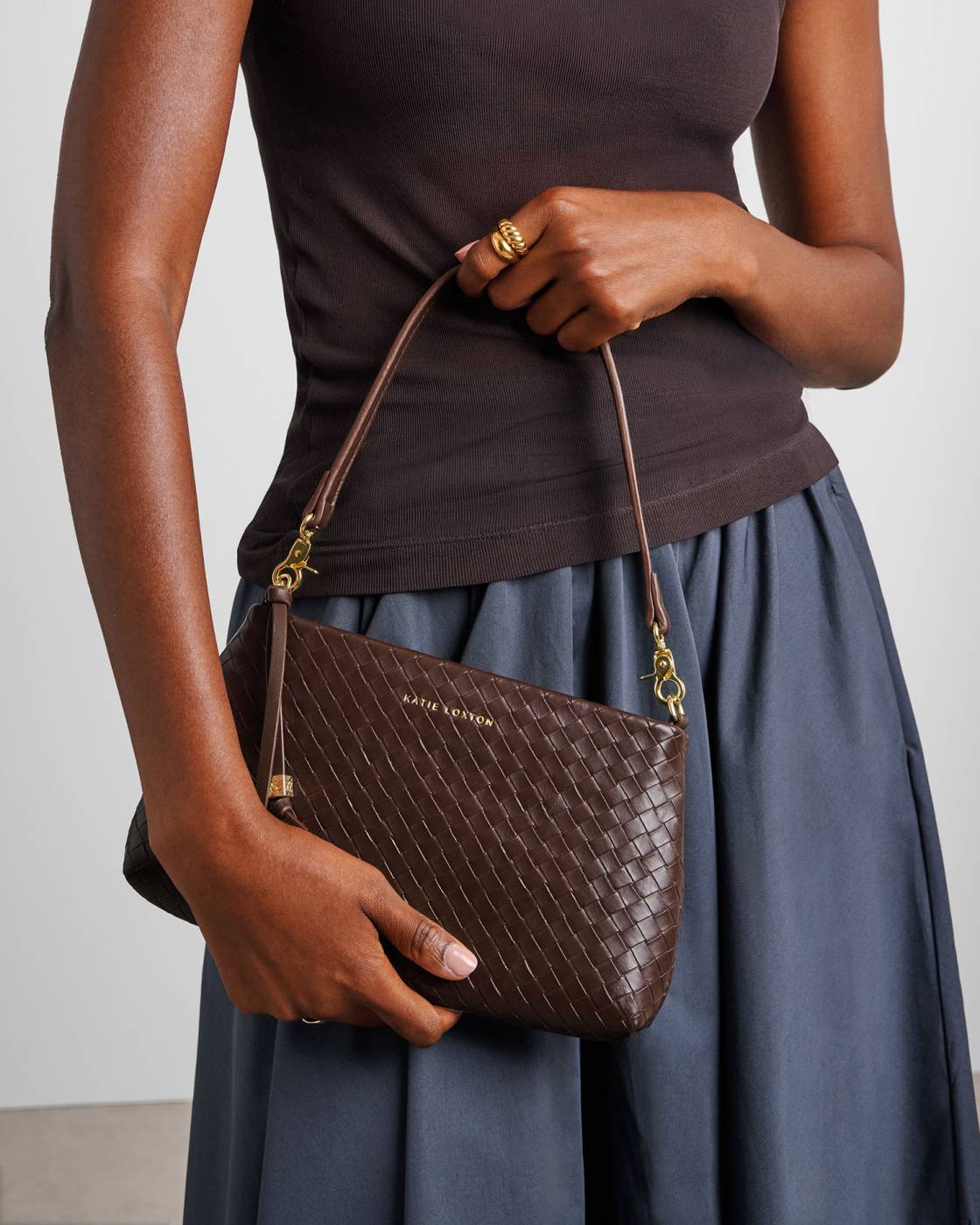 A model holds a brown woven leather shoulder bag with gold hardware and a tassel charm. The model is wearing a dark brown top and a slate blue skirt against a light gray background.