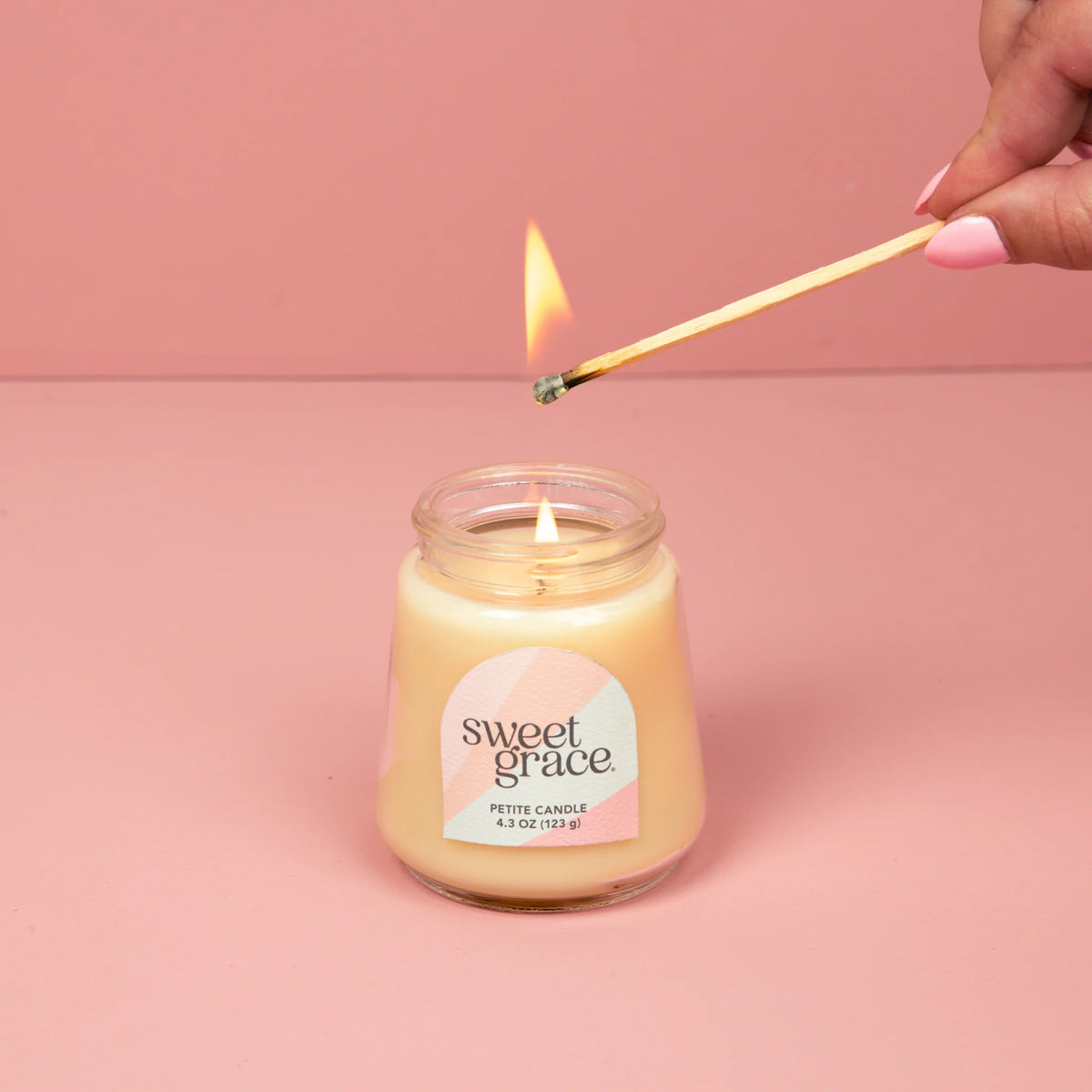 Person lighting a 'sweet grace' candle with a match on a pink background