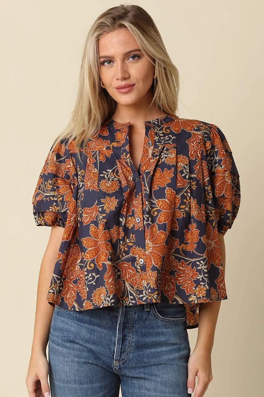 A woman wearing a short-sleeve blouse with a navy blue base and an orange floral pattern, paired with blue jeans, against a solid beige background.