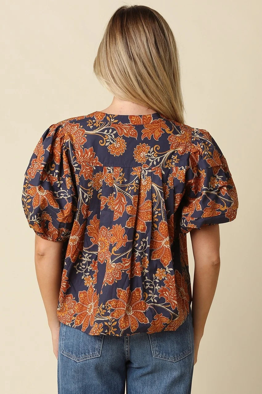 A woman viewed from the back wearing a short puff-sleeve blouse with an orange and brown floral print on a navy blue background, paired with blue jeans, against a solid beige studio backdrop.