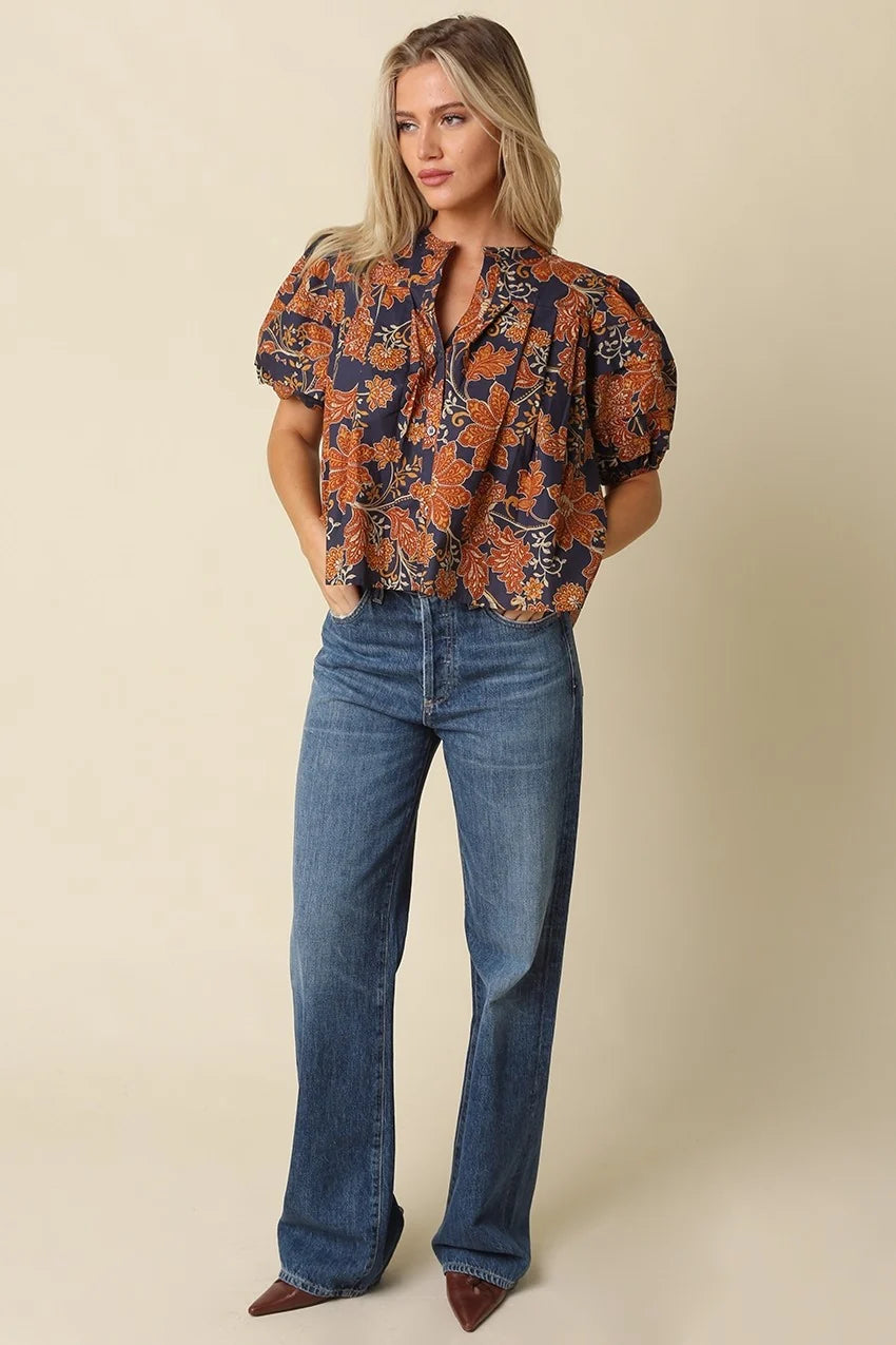 A woman modeling a short-sleeve blouse with a navy blue and rust-orange floral pattern, paired with blue jeans and brown pointed-toe heels, against a solid beige studio background.