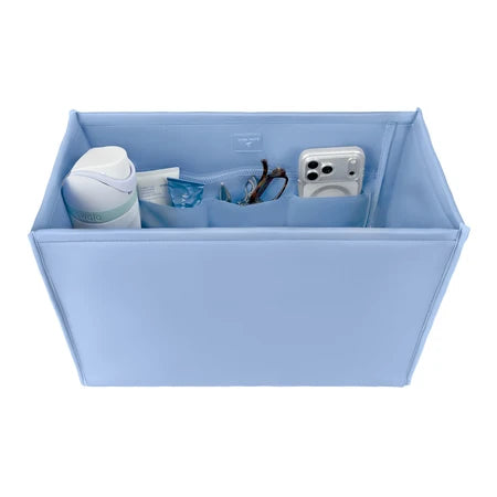 A light blue fabric organizer box with multiple interior pockets, shown holding various small items like a white bottle, a clear pouch, and a white camera. The box is photographed on a clean white background.