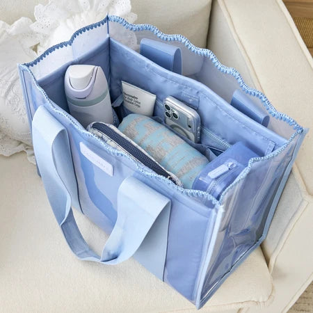 A light blue translucent tote bag with matching fabric handles, shown open and filled with toiletries and accessories on a cream-colored sofa.