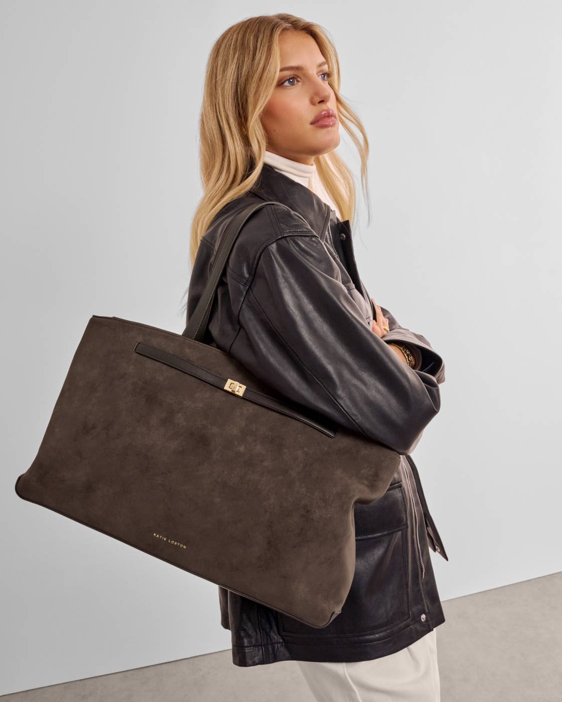 A woman wearing a black leather jacket and cream turtleneck carries a large dark brown suede tote bag with a black leather strap and gold hardware.