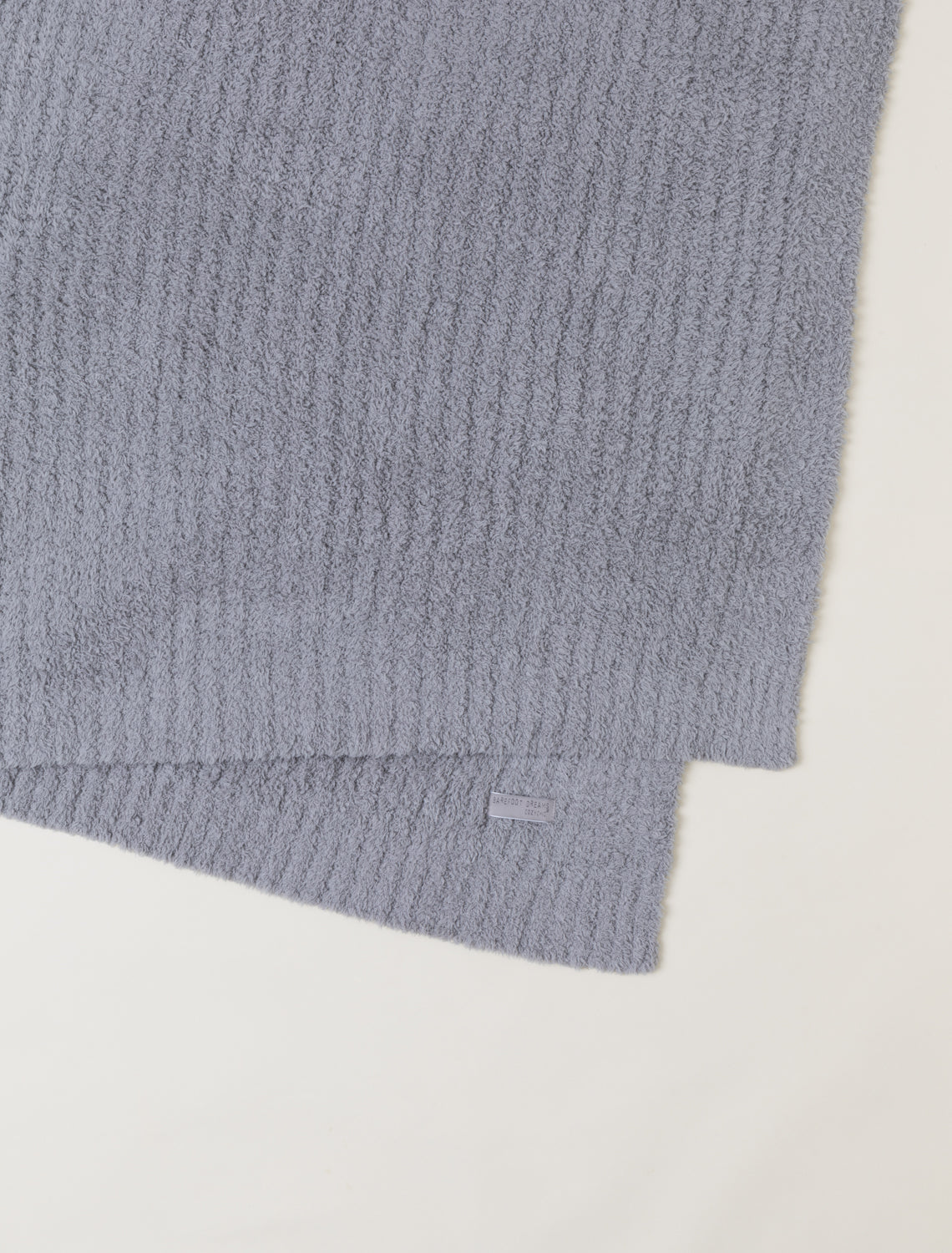 A close-up of a folded ribbed knit fabric in a muted dusty lavender-gray color, showing the soft texture and a small matching fabric tag, on a plain off-white background.
