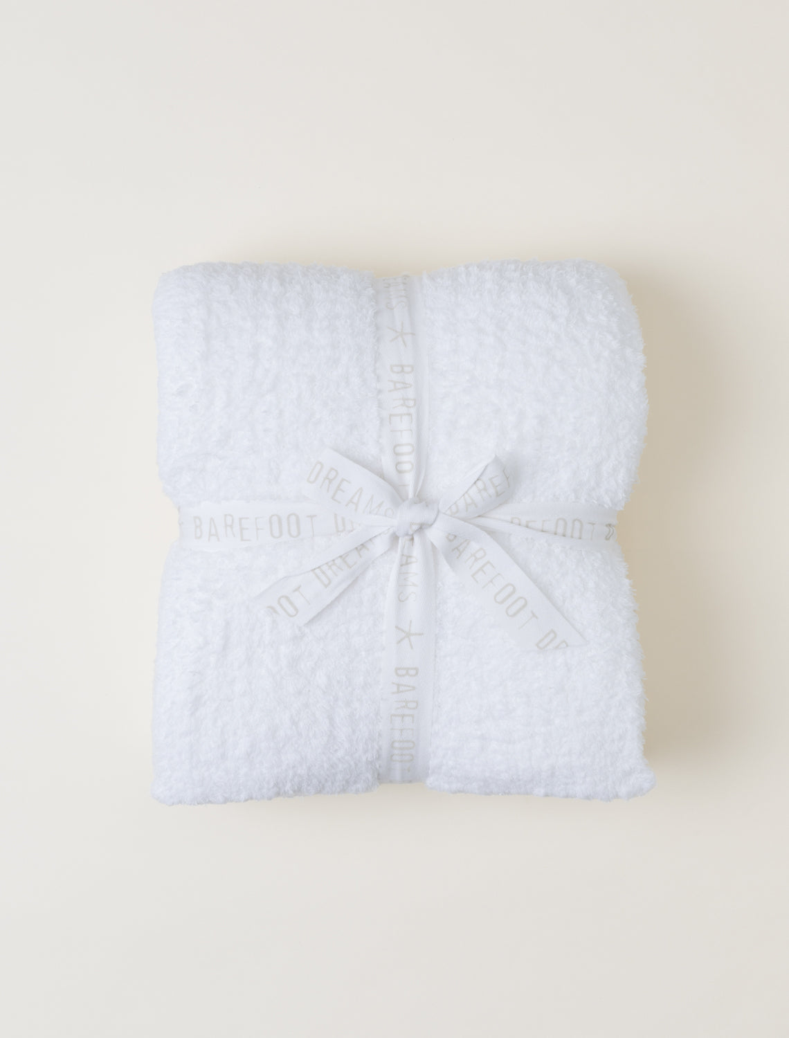 A folded white blanket wrapped with a white ribbon printed with the brand name 'BAREFOOT', tied in a bow, on a light beige background.