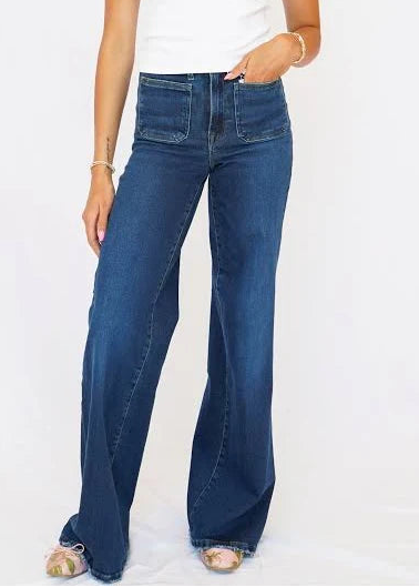 A woman modeling a pair of high-waisted, wide-leg blue jeans with a white ribbed tank top, standing against a plain white studio background.