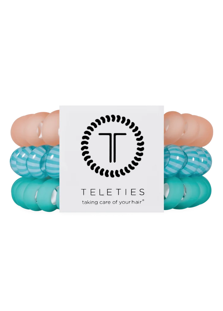 Three Teleties hair elastic bands in peach, light blue, and turquoise, stacked behind a white brand card on a solid white background.
