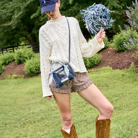 A person outdoors on grass wearing a cream cable-knit sweater, brown gingham shorts, brown cowboy boots, a navy cap, and a blue crossbody bag, while holding a blue and white pom-pom.