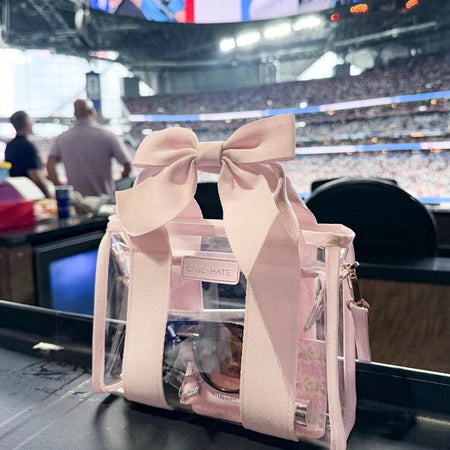 A pink and clear tote bag with a large pink bow on a stadium table during a game, featuring a visible Rose Nate brand label.