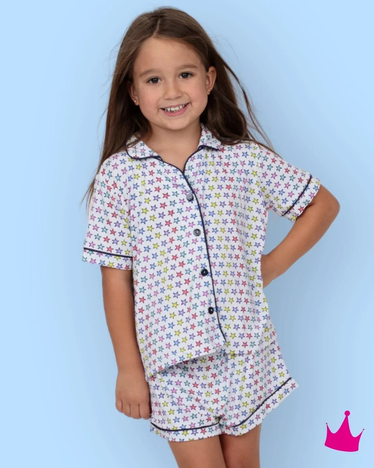 A young girl with long brown hair smiling while wearing a two-piece pajama set with a colorful star pattern on a white background, set against a solid light blue backdrop.