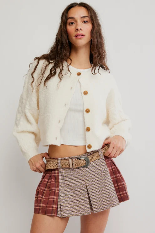 A model wearing a cream textured cropped cardigan with brown buttons, a white crop top, and a pleated mini skirt with a red and beige plaid pattern and a tan belt.