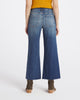 Rear view of a model wearing blue wide-leg denim jeans with a frayed hem, paired with a mustard yellow ribbed top and brown ankle boots, against a light gray studio background.