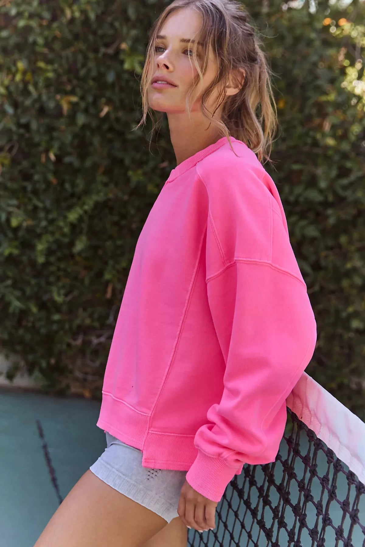 A woman wearing a bright pink crewneck sweatshirt and light gray denim shorts, leaning against a tennis net with a green hedge in the background.