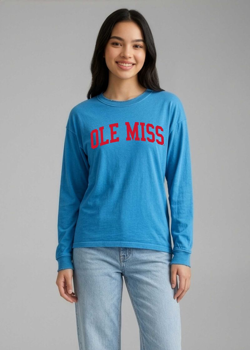 A woman modeling a bright blue long-sleeve crewneck sweatshirt with 'OLE MISS' printed in red across the chest, paired with light-wash jeans against a solid gray studio background.